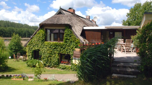Una casa con techo de paja y paredes cubiertas de hiedra, un patio con mobiliario exterior y un jardín con flores y plantas.