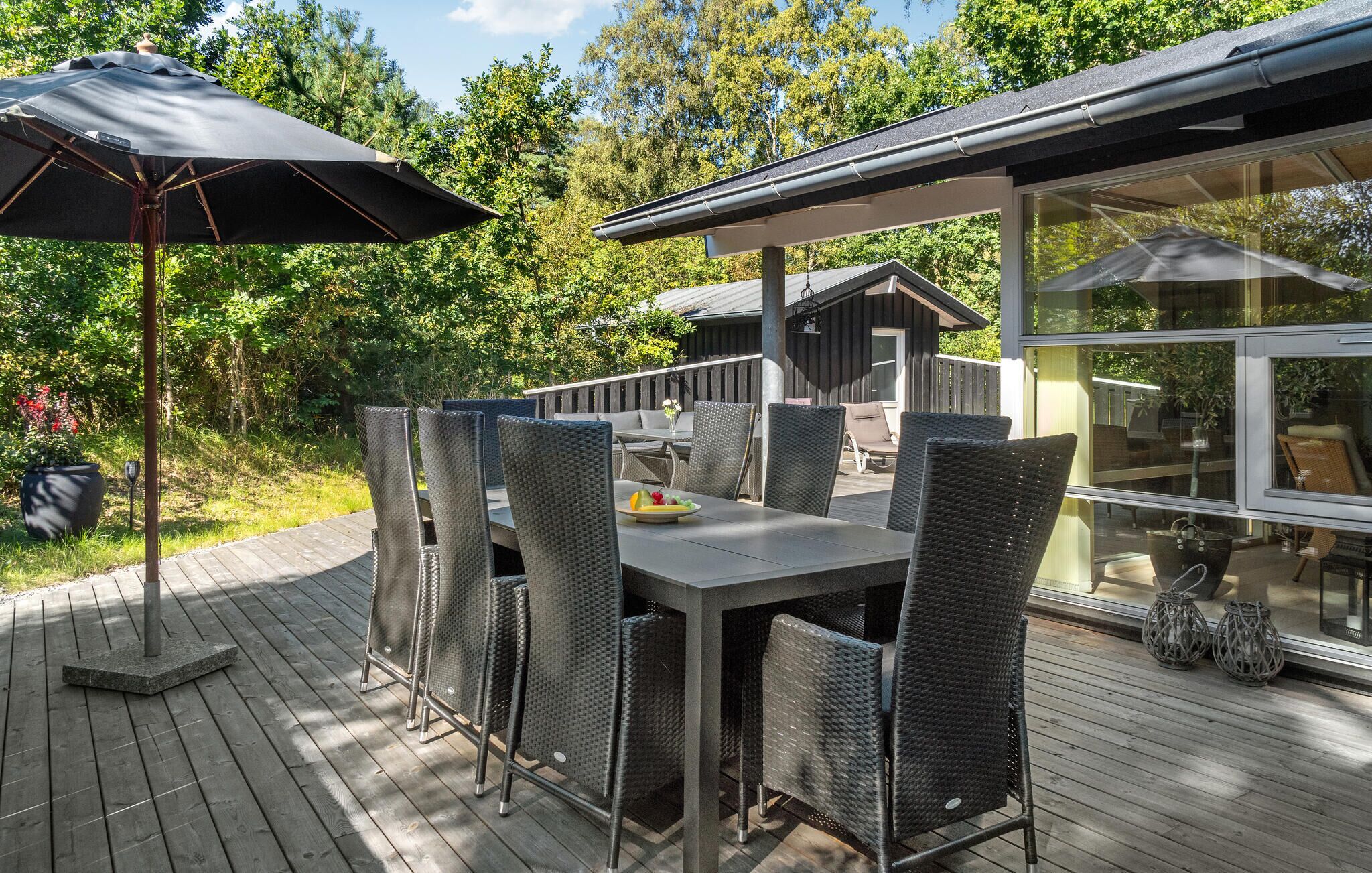 Eine Terrass mit Esstisch und Stühlen, einem großen Sonnenschirm und einem gläsernen Anbau mit Sitzgelegenheit.