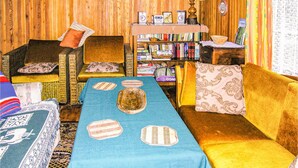 Ein gemütliches Wohnzimmer mit einem blauen Sofa, einem gelben Sofa und einem Korbstuhl. Ein Bücherregal ist mit Büchern gefüllt und ein kleiner Tisch trägt eine Schale und einen Bilderrahmen.