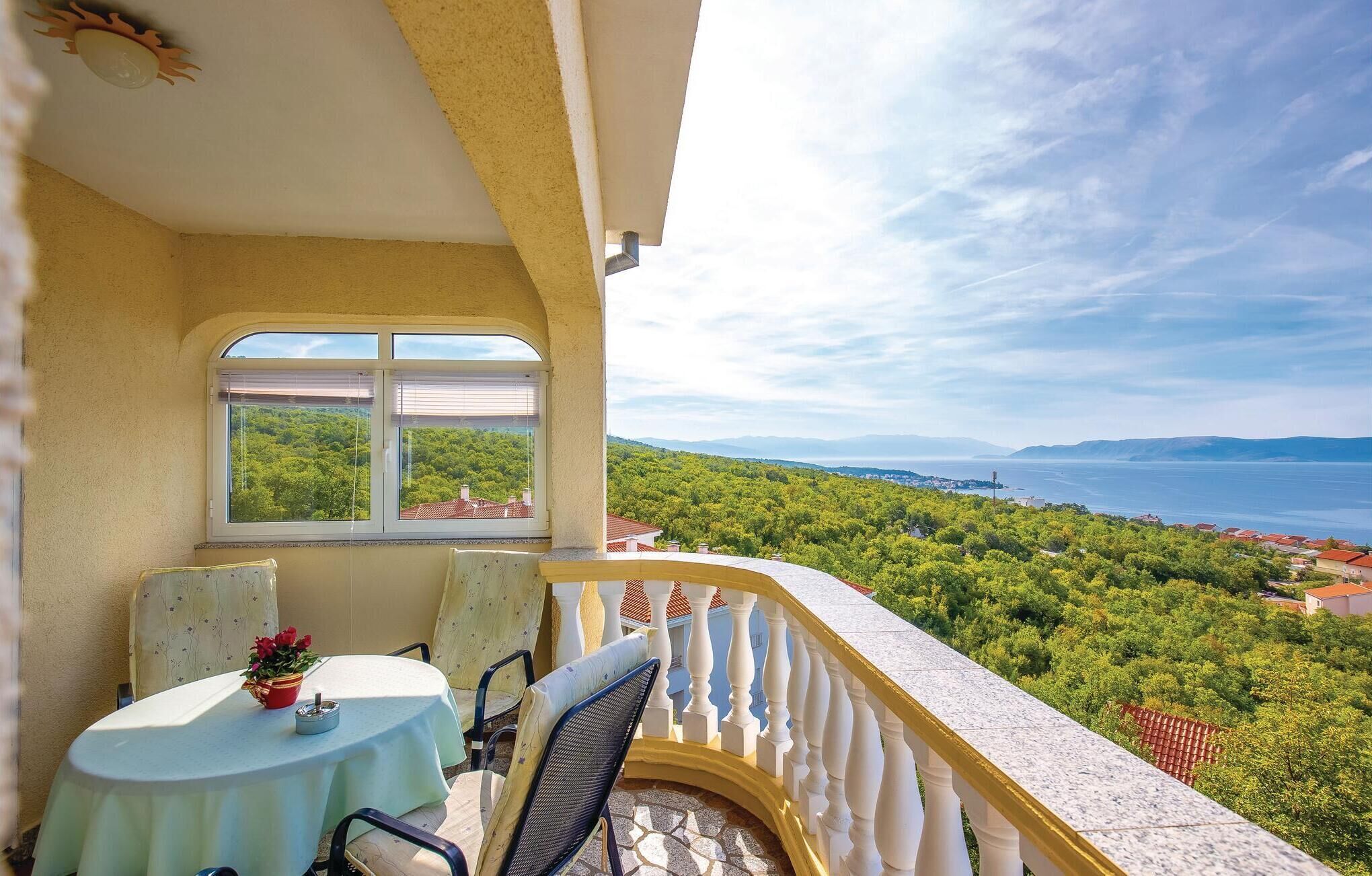 Un balcon avec une table et des chaises, une fenêtre donnant sur un paysage verdoyant et une étendue d’eau visible au loin.