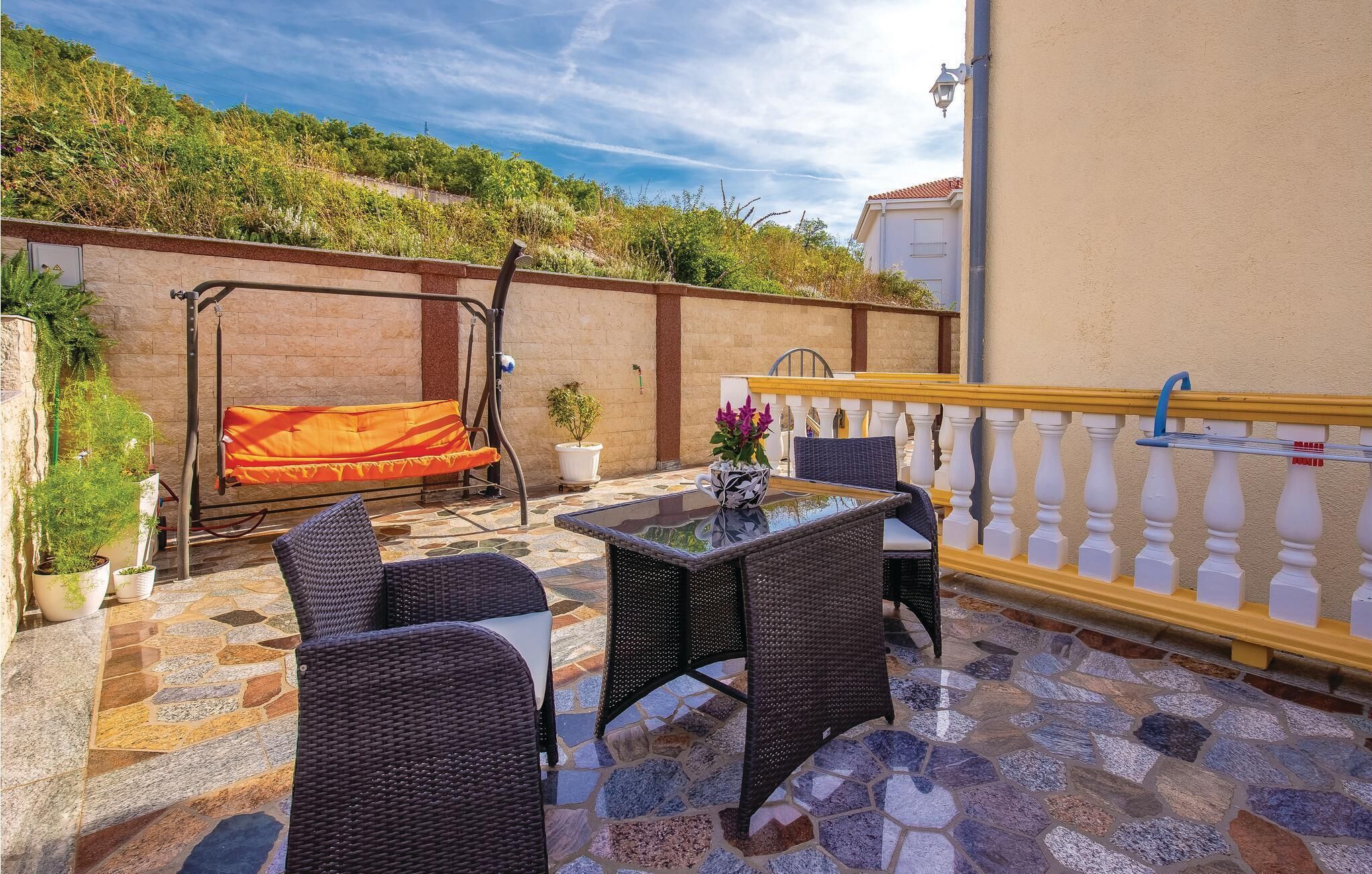 Un patio al aire libre con mobiliario de mimbre, una mesa de vidrio y una barandilla amarilla.