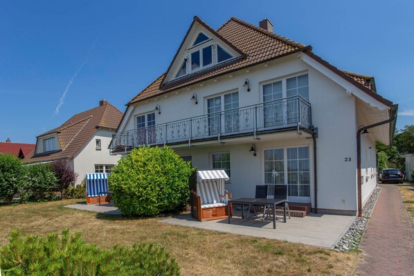 Une maison à deux étages avec un balcon, une terrasse dotée d’une table et de chaises, et un petit jardin.