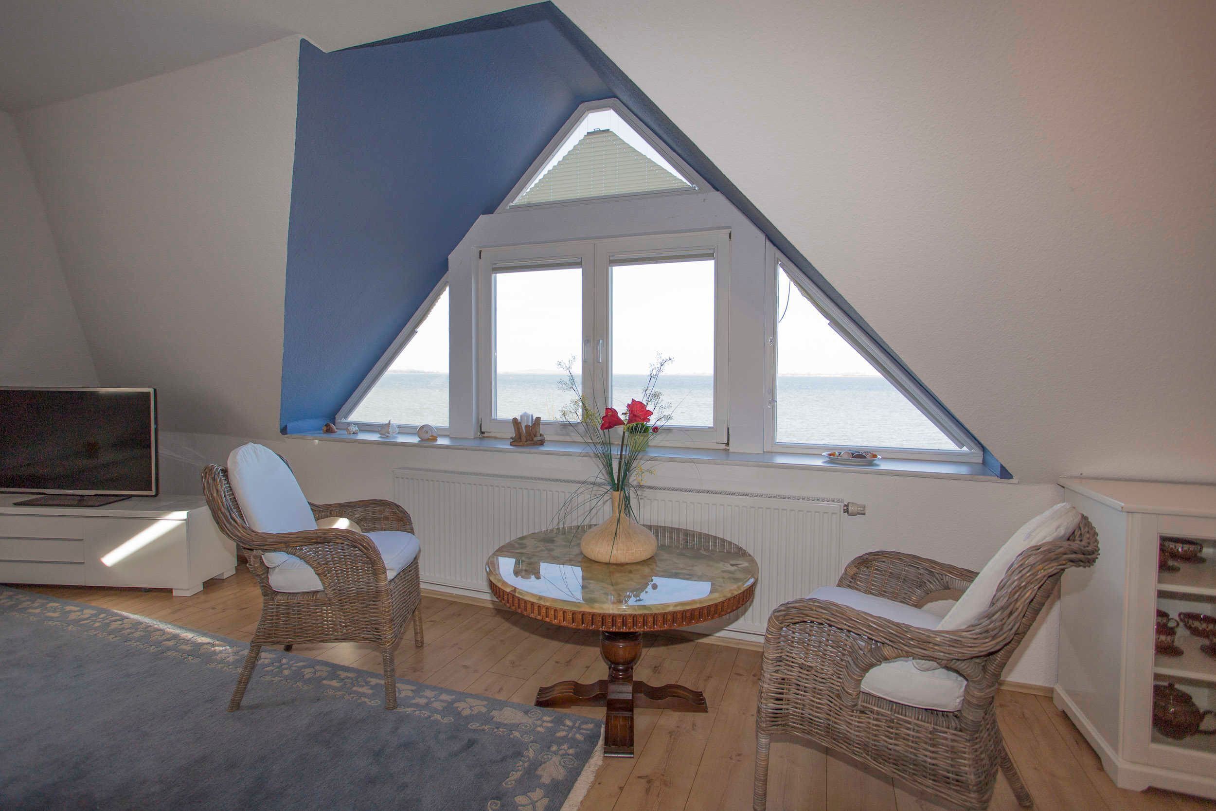 Ein Zimmer mit schrägen Wänden, einem großen Fenster mit Meerblick, Korbmöbeln, einem runden Tisch mit einer Vase und einem Fernseher links.