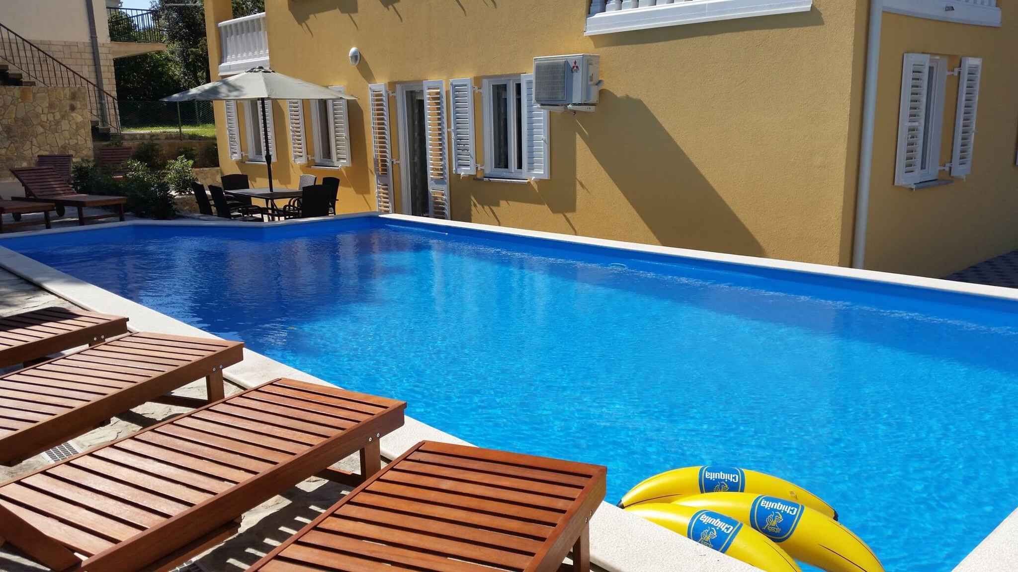Una piscina amb gandules de fusta i una taula, envoltada d'un edifici groc amb persianes blanques.
