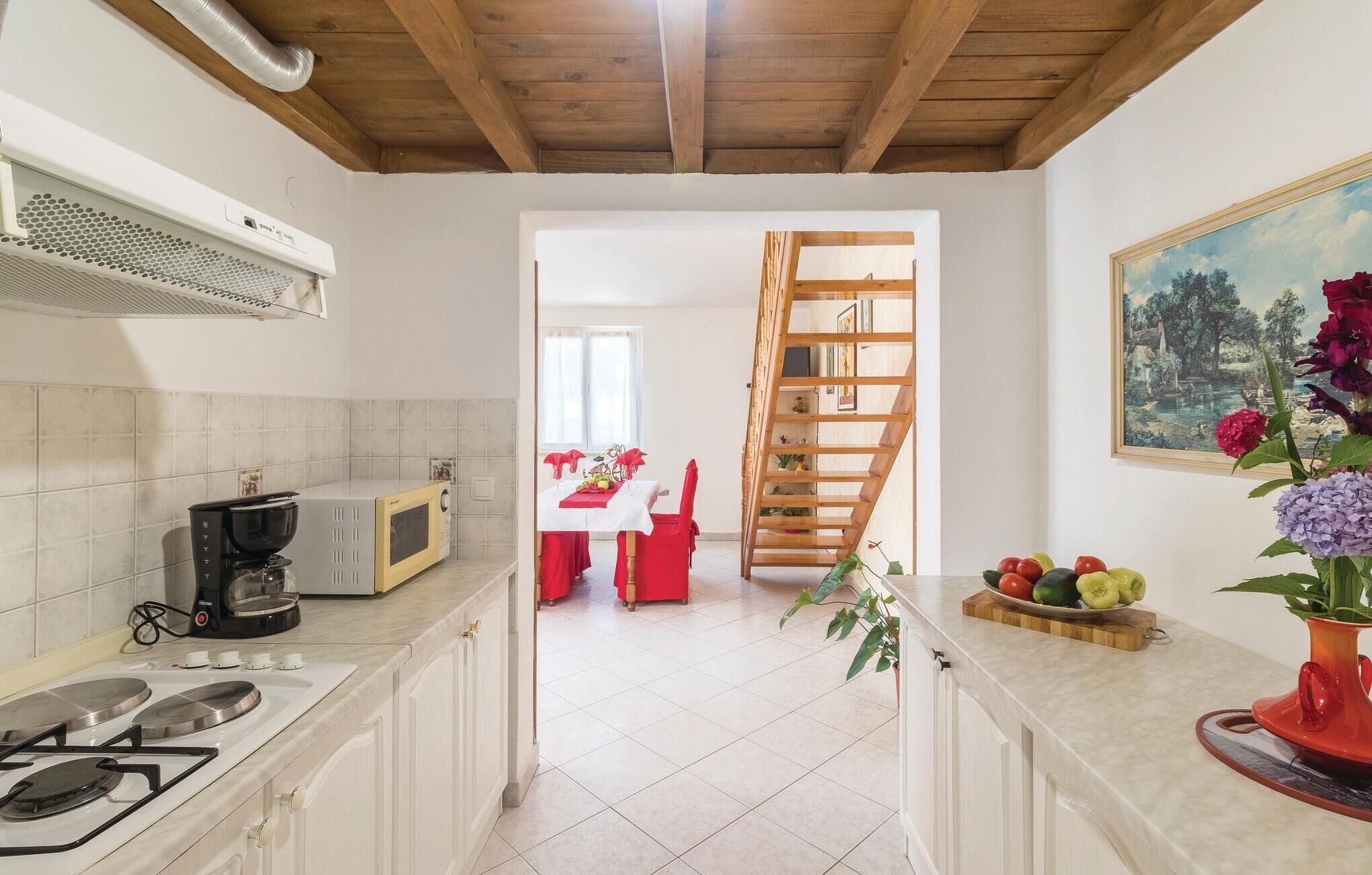 Una cocina con estufa a gas, microondas y cafetera. Hay un comedor con una mesa y sillas rojas, y una escalera de madera que lleva a otra habitación.