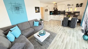 Una sala de estar moderna con un sofá gris y cojines azules, una mesa de centro blanca y una cocina con armarios blancos y encimeras negras.