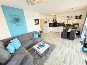 A modern living room with a grey sofa and blue cushions, a white coffee table, and a kitchen area with white cabinets and black countertops.