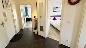 Un pasillo de hotel con diseño moderno, que presenta un suelo de baldosas oscuras, una puerta blanca con espejo, un cuadro enmarcado, un teléfono y una planta decorativa.