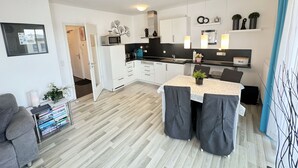 Una cocina moderna con armarios blancos, una zona de comedor con mesa y sillas, y un pequeño rincón de estar con un sofá y libros.