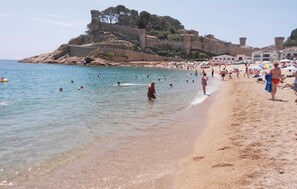 Een strand met helder water, mensen die zwemmen en langs het strand wandelen, en een historisch fort dat uitkijkt over het strand.