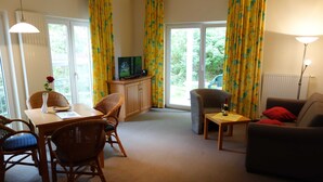 A living room with a dining area, a television, wicker furniture, and large windows with curtains.