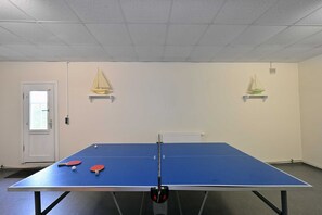 A ping pong table with two paddles and a ball, a white door, and two small sailboat decorations on shelves.