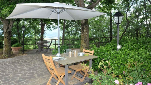 An outdoor patio with a table and chairs, a large umbrella, and a view of the surrounding greenery.