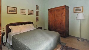 A bedroom with a bed, a wooden wardrobe, a floor lamp, and framed pictures on the wall.