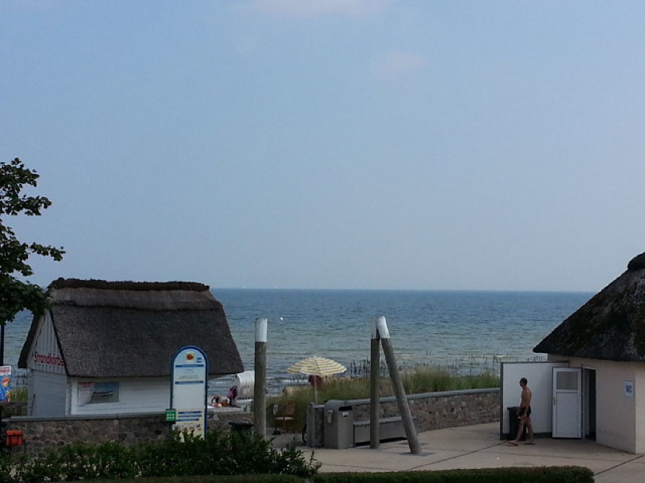 A coastal view with a thatched-roof building, a signboard, and a person near a small structure.
