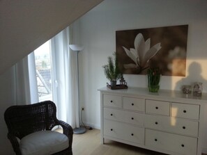 A room with a white dresser, a wicker chair, a large window, and a floor lamp.