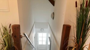 Una escalera conduce a una habitación con una puerta blanca, barandas de madera y plantas en macetas a ambos lados.