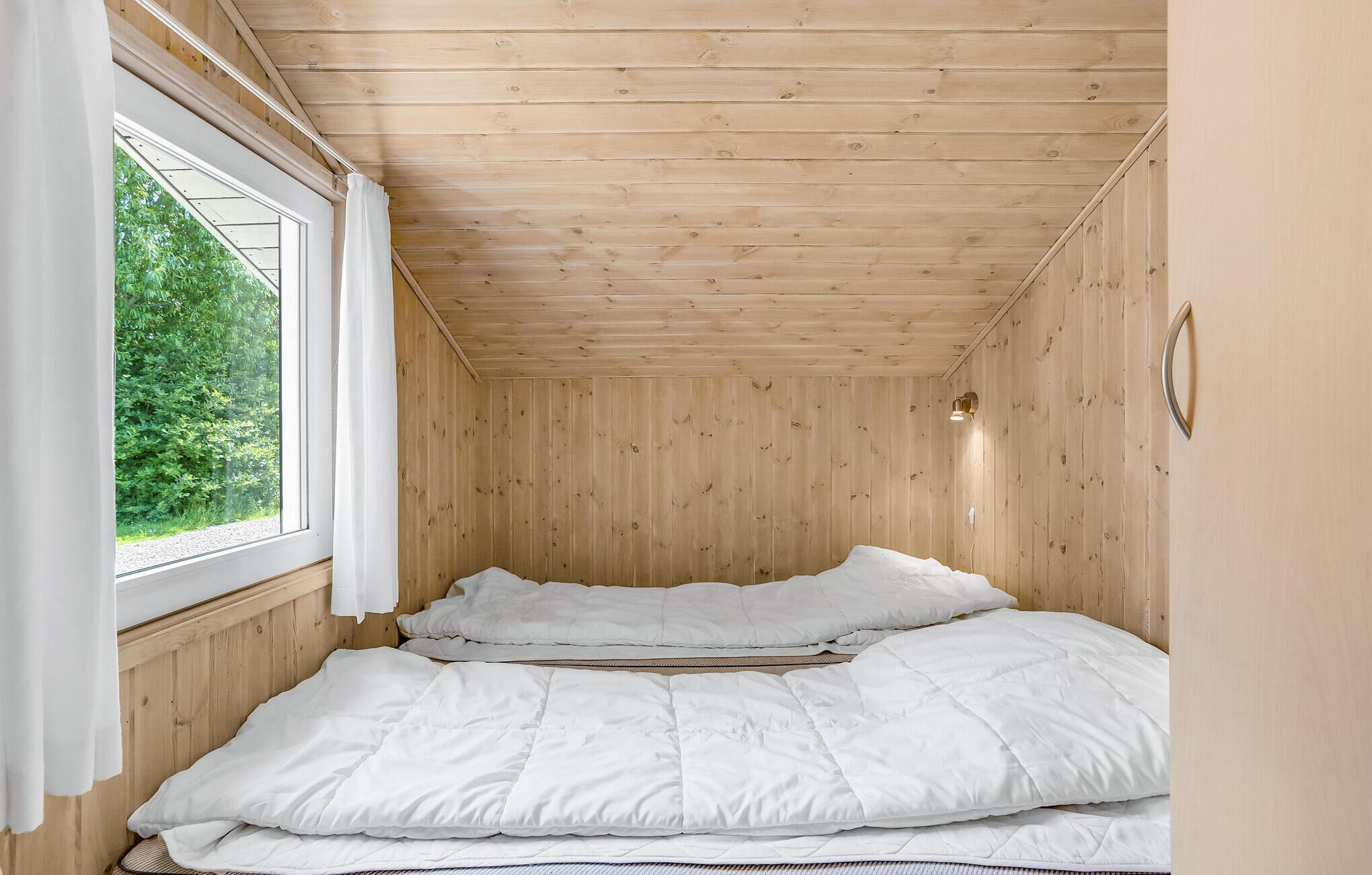 Una habitación pequeña con paneles de madera, una cama, un ventanal con cortinas y una lámpara fijada a la pared.