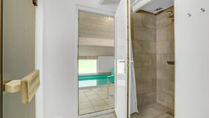 Ein Badezimmer mit Glastür zu einem Balkon, einer Duschkabine mit beigen Fliesen und einem Fenster mit Jalousien.