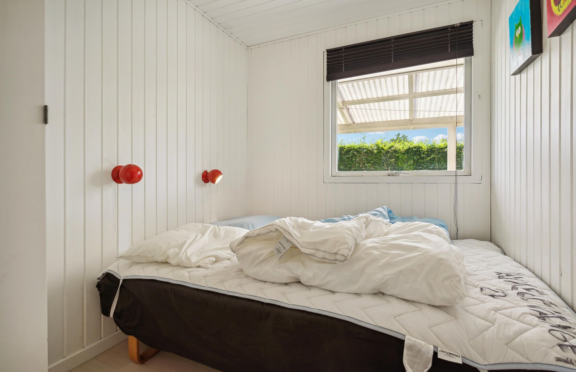 A bedroom with a bed, a window with a view of greenery, and two red wall-mounted lights.