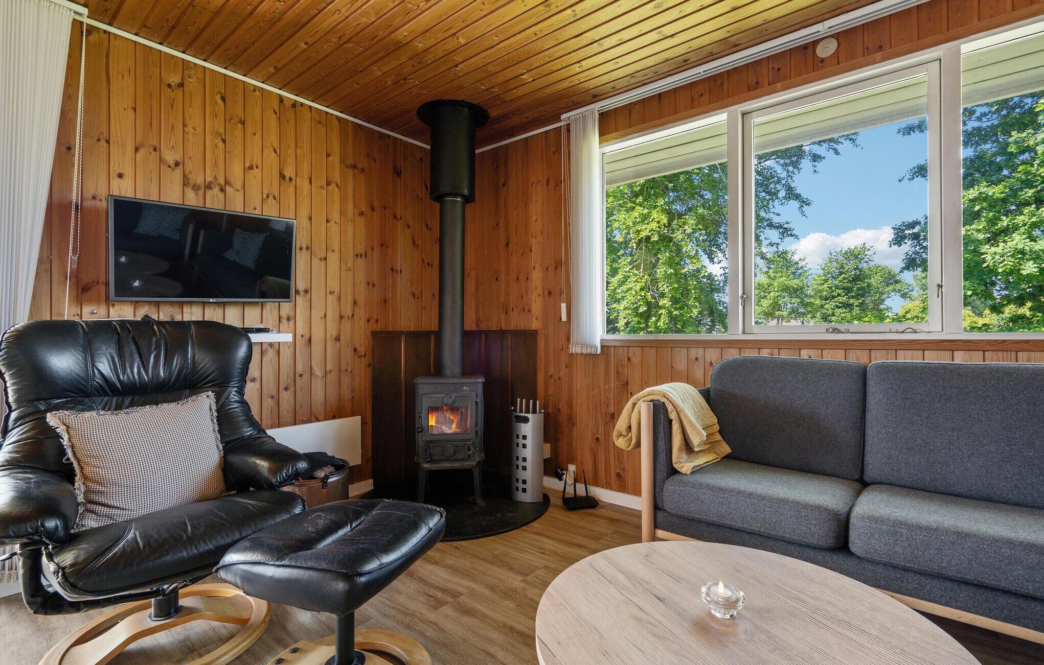 A cozy living room with a leather chair, a gray sofa, a wooden table, a TV mounted on the wall, and a wood-burning stove.