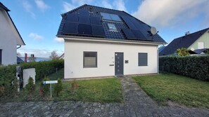 Uma casa residencial com painéis solares, um pequeno jardim frontal e um acesso pavimentado.