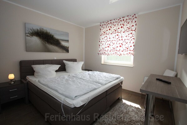 Une chambre à coucher avec un lit, une table de chevet, une lampe, une fenêtre avec des rideaux à motifs et un tableau au mur.