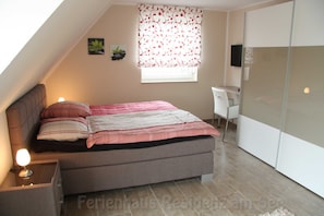 Een slaapkamer met een bed, nachtkastje, lamp, raam met bloemen gordijnen en een bureau met stoel.