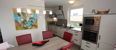 Une cuisine moderne avec un coin repas, agrémentée d’un tableau, de chaises rouges et d’un four encastré.