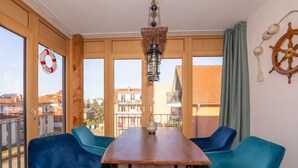 Un coin repas avec une table en bois, deux chaises bleues, une lanterne suspendue et une vue sur les bâtiments à travers de grandes fenêtres.