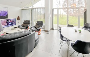 Ein modernes Wohnzimmer mit Fernseher, Ledersofas, einem Esstisch und großen Fenstern.