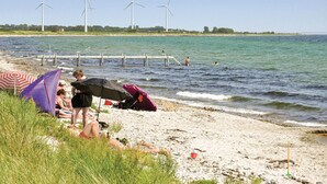 Een strand met zonnebaders, een pier met windturbines op de achtergrond en een heldere lucht.