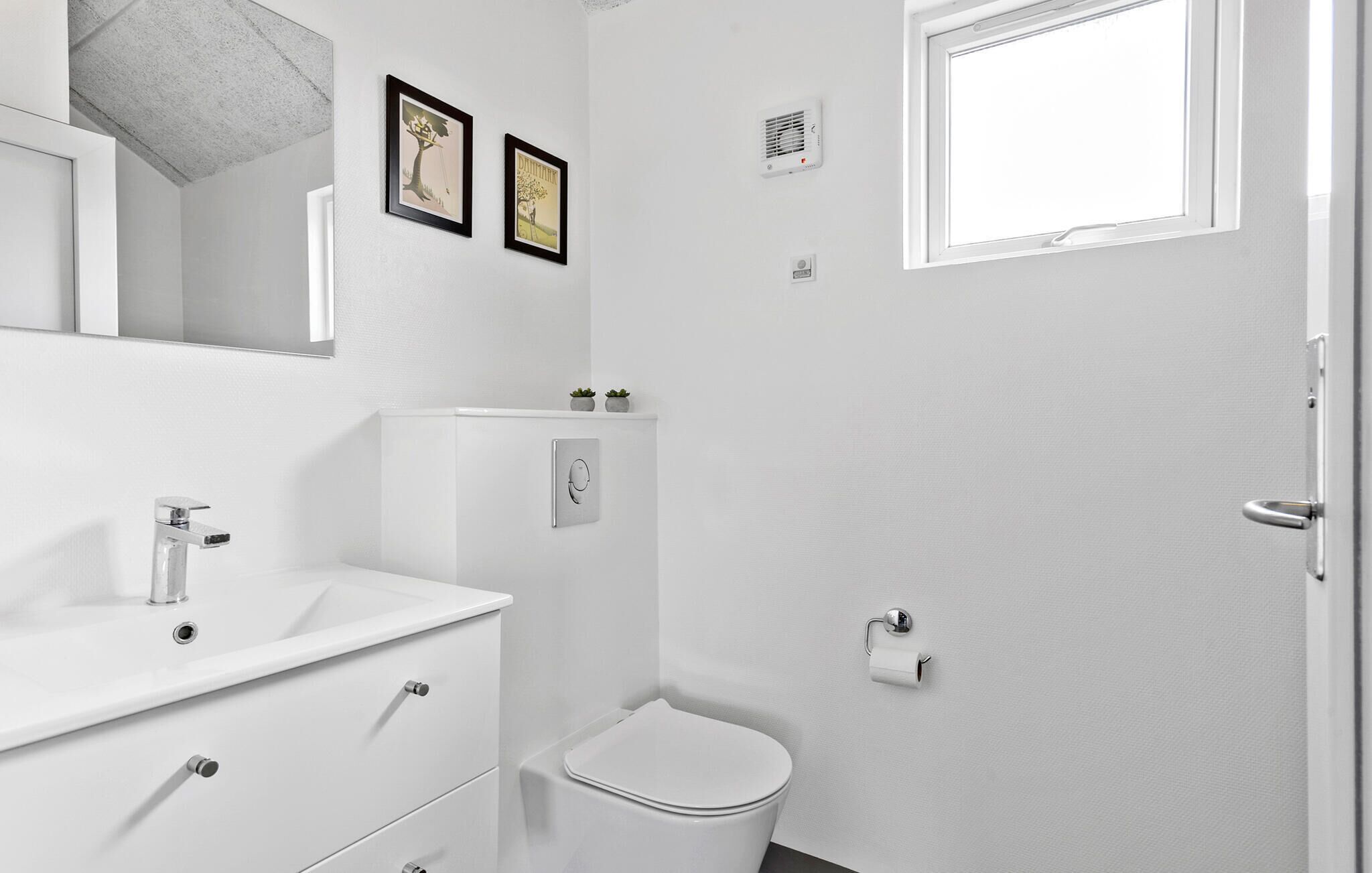 Une salle de bain moderne avec un lavabo blanc, un miroir et des toilettes. On y trouve deux tableaux encadrés au mur, une petite fenêtre et un système de ventilation.