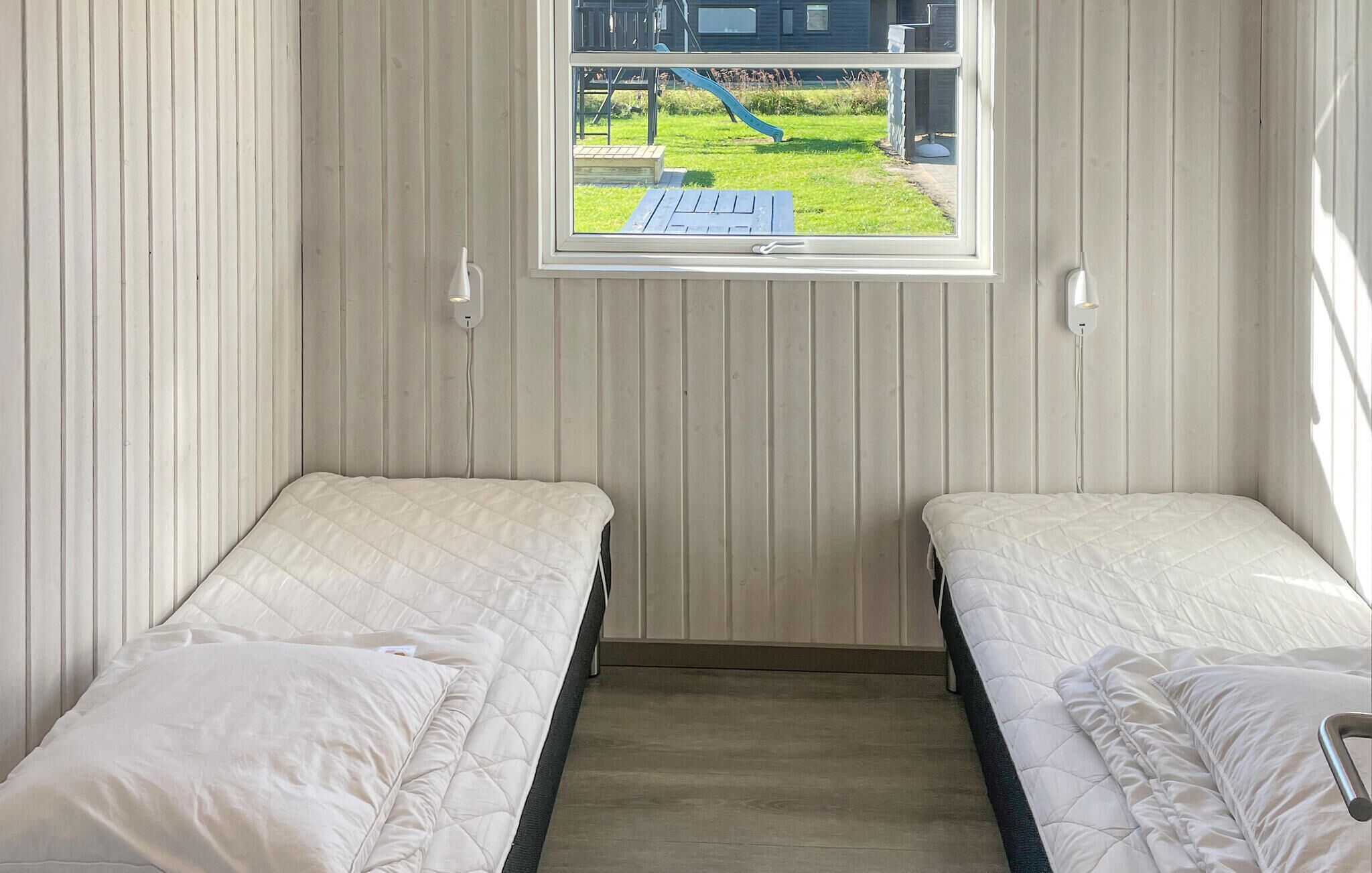 A room with two single beds, a window with a view of a playground, and a wall-mounted light fixture.