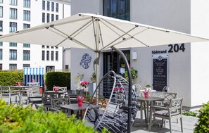 Een open café met tafels en stoelen, een grote parasol en een gebouw met nummer 204.