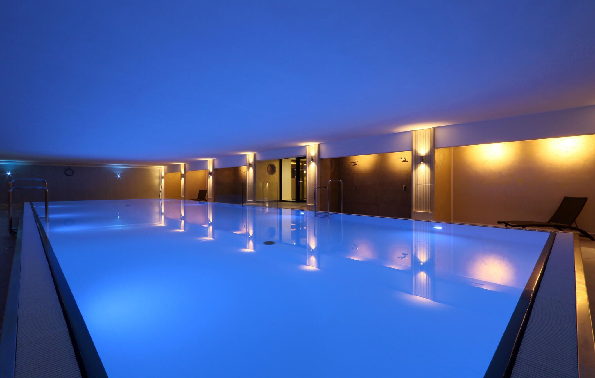 Ein großes Hallenbad mit Glastrennwand, warmer Beleuchtung und einigen Liegestühlen.