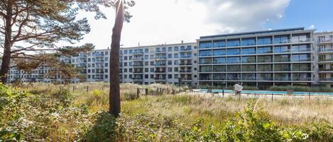 A modern apartment building with multiple balconies, a swimming pool, and a grassy area with trees.