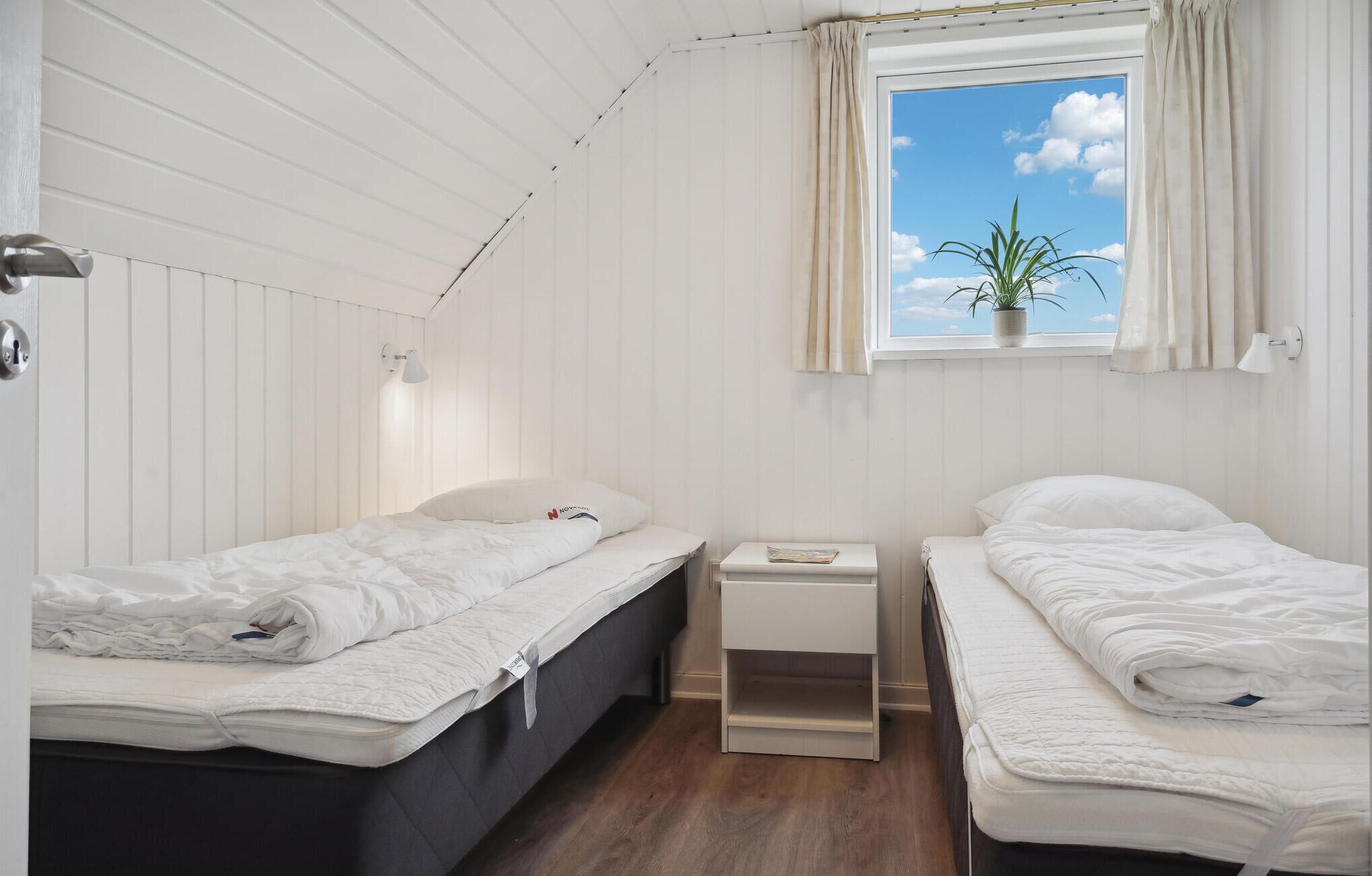 A small, clean room with two single beds, a bedside table, a window with a view of the sky, and a potted plant.