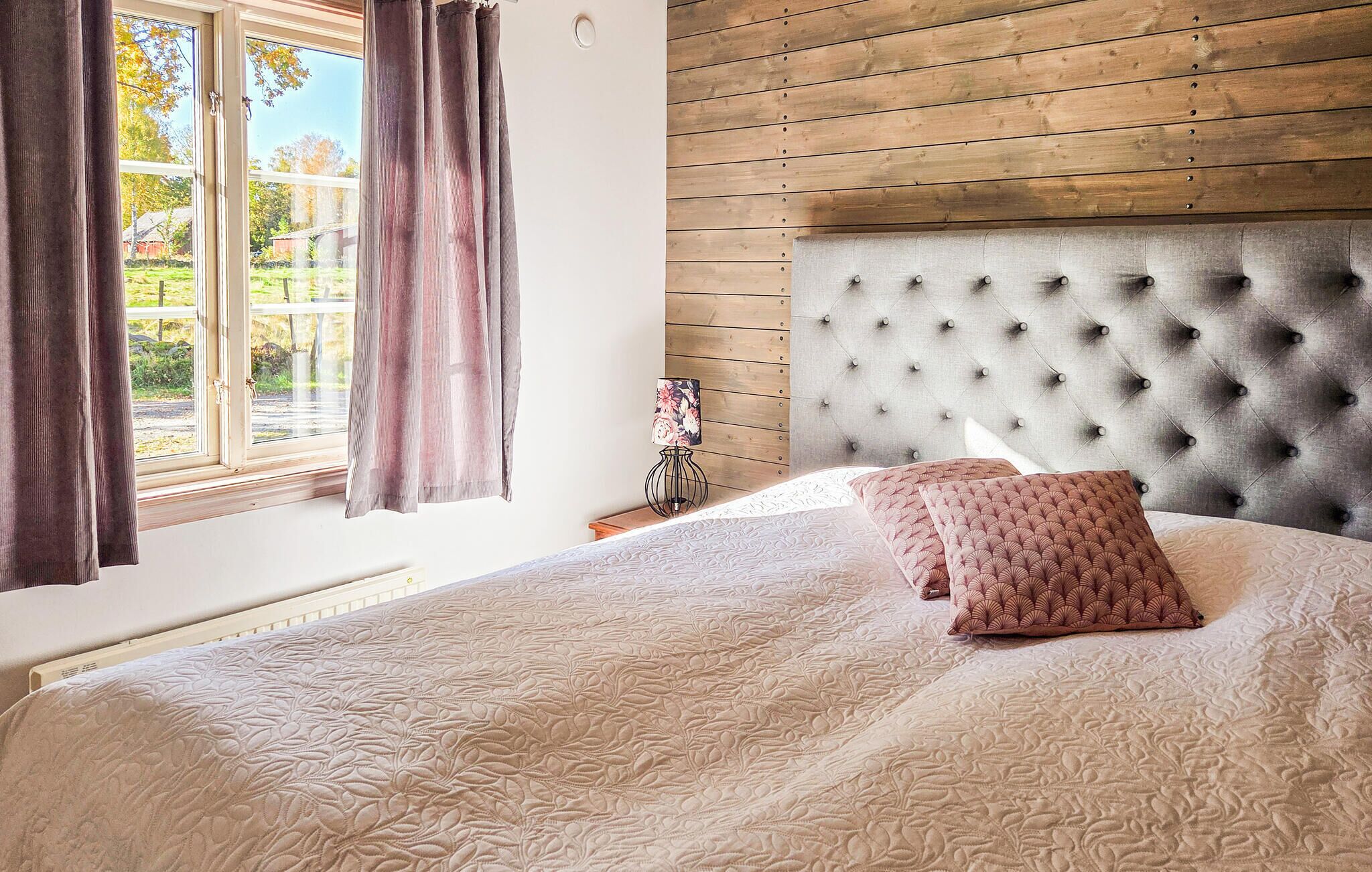 Une chambre à coucher avec une tête de lit capitonnée, un lit avec une literie à motifs, une fenêtre avec des rideaux et une table de chevet avec une lampe.