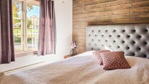 Una camera da letto con una testiera imbottita, un letto con biancheria a motivi, una finestra con tende e un comodino con una lampada.