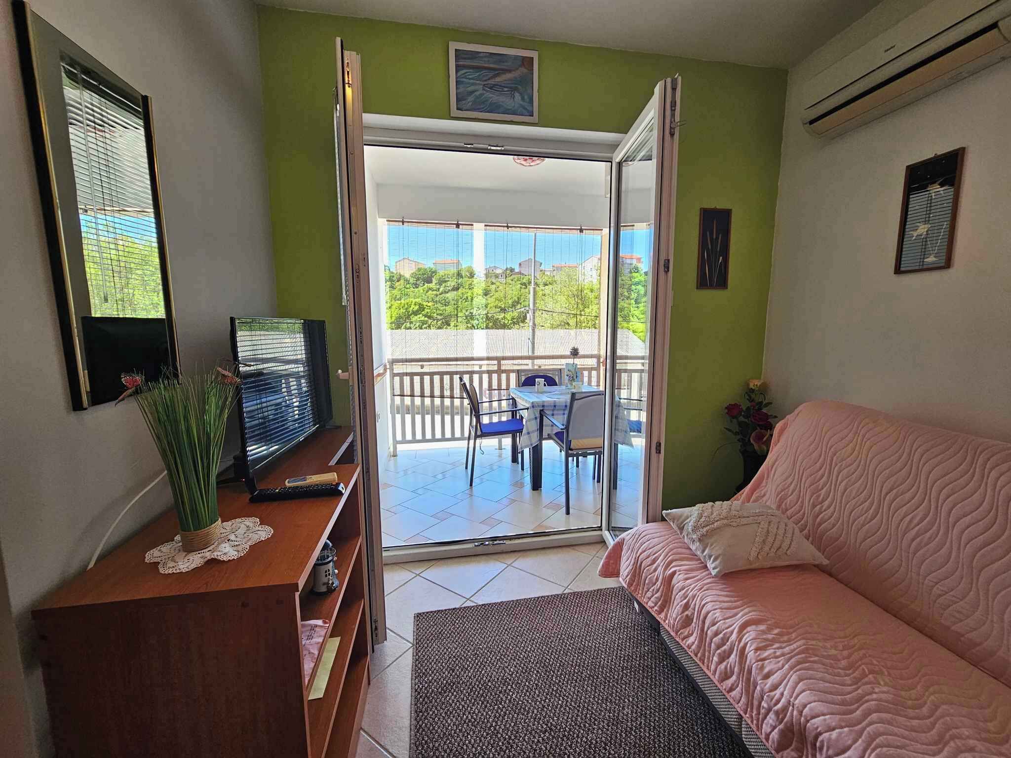 A room with a sofa, a TV on a wooden stand, a table with chairs, and a balcony with a view of greenery and buildings.