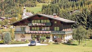 A two-story chalet with a balcony, surrounded by a well-maintained garden and a mountainous backdrop.