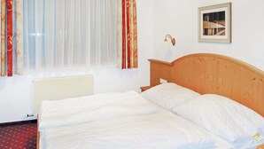 A room with a wooden bed, a radiator, a window with sheer curtains, a wall-mounted lamp, and a framed picture.
