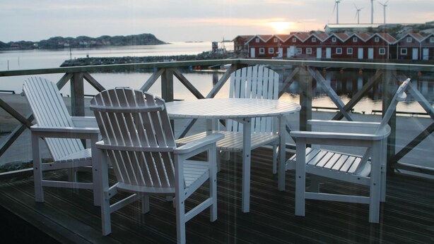 Een houten terras met een ronde tafel en twee Adirondack-stoelen, met uitzicht op een waterpartij en een rij rode gebouwen met windturbines op de achtergrond.