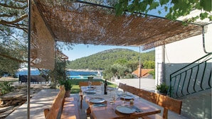Un espace de restauration en plein air avec une table dressée pour quatre personnes, une pergola au-dessus et une vue sur la mer et les collines en arrière-plan.