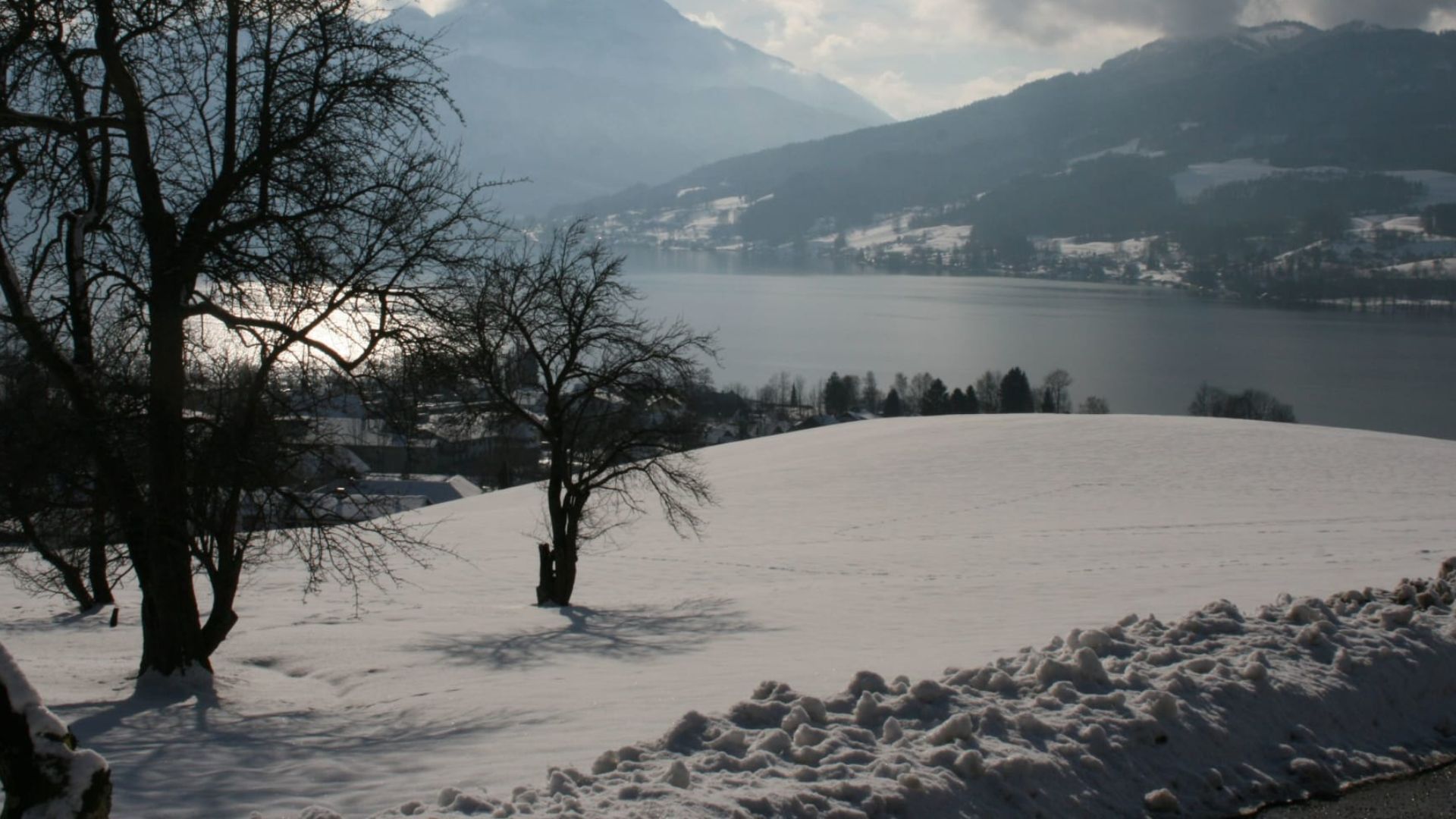 A snow-covered landscape with a lake, mountains, and leafless trees.