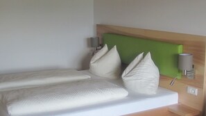 Una habitación de hotel con cama, almohadas, cabecera verde, lámpara de pared y toma de corriente.