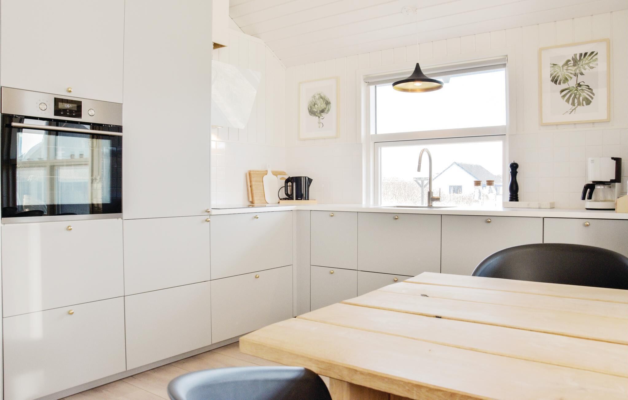 Une cuisine moderne avec des meubles blancs, un four encastré, une table à manger en bois et une fenêtre donnant sur une maison.