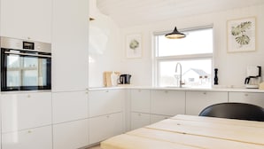 A modern kitchen with white cabinetry, a built-in oven, a wooden dining table, and a window with a view of a house.