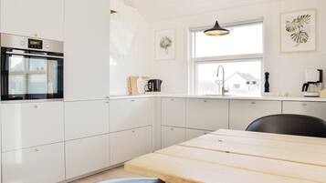 Et moderne køkken med hvide skabe, en indbygget ovn, et træspisebord og et vindue med udsigt til et hus.